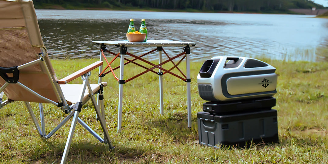 The Mark 3 portable air conditioner sits near a lake, placed on a storage box next to a camping chair and a table with drinks and snacks, providing a comfortable cooling solution for outdoor relaxation.